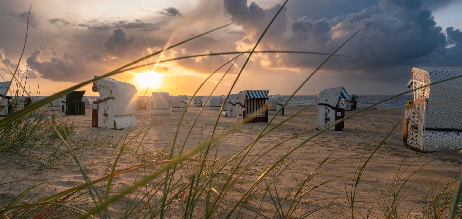Hübscher Sonnenuntergang über einem Strand mit Strandkörben und grünem Gras im Vordergrund.